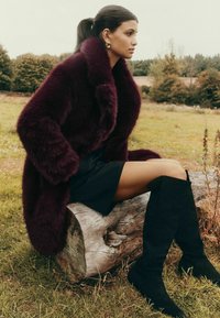 Burgundijas mākslīgā kažoka mēteli ar lielu apkakli, apvienots ar melnu mini svārku un melniem zamša zābakiem līdz ceļiem. Modelis sēž uz laukakmens.