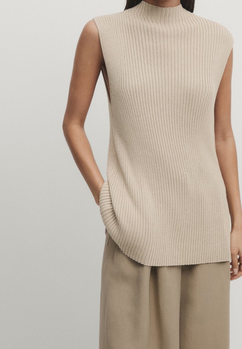 Femme portant un top beige en maille côtelée sans manches et un pantalon beige ample, une main dans la poche, sur un fond uni.