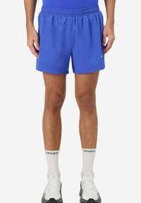 Pantaloni sportivi blu con elastico in vita, caratterizzati da una texture liscia e un accento con logo. Abbinati a calzini bianchi e scarpe da ginnastica chiare.