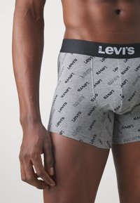 Graue Baumwoll-Boxershorts mit einem schwarzen Bund, auf dem wiederholt das Levi's-Logo in verschiedenen Ausrichtungen zu sehen ist. Nahaufnahme des Kleidungsstücks und des Oberschenkels.