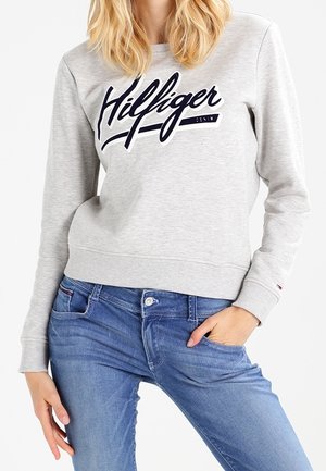 Vrouw in een lichtgrijze trui met het logo "Hilfiger" en een blauwe spijkerbroek, met één hand in de zak en de andere ontspannen langs het lichaam.