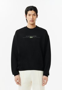 Sudadera negra hecha de mezcla de algodón, con cuello redondo, mangas largas y logo bordado 'LACOSTE' en verde en la parte frontal.