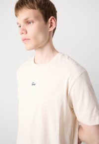 Beige bomulls T-skjorte med rund hals og korte ermer, med diskret brodert logo på brystet. Glatt tekstur, tettsittende design.