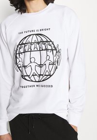 Vit sweatshirt med svart grafisk tryck av en glob och en pappersdockkedja, text ovan "FRAMTIDEN ÄR LJUS", nedan "TILLSAMMANS NÅR VI FRAMGÅNG".