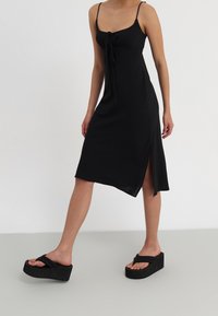 Vestido negro sin mangas con un detalle de lazo en el frente, abertura lateral y tejido texturizado, combinado con sandalias de plataforma negras gruesas.