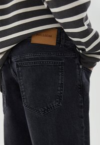 Jeans de mezclilla negra con un corte recto, que presentan un parche de cuero marrón de Pull&Bear y costuras en contraste. Se pueden ver los bolsillos laterales y traseros.