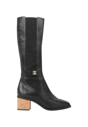 Botte haute noire en cuir, avec un talon bloc en liège et un petit emblème de logo argenté rond sur le côté près de la cheville.