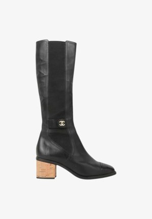Botte haute noire en cuir, avec un talon bloc en liège et un petit emblème de logo argenté rond sur le côté près de la cheville.