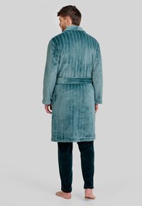 Bata de baño de felpa turquesa con rayas verticales, un cinturón en la cintura y una textura suave. Combinada con pantalones de pijama oscuros verdes y suaves.