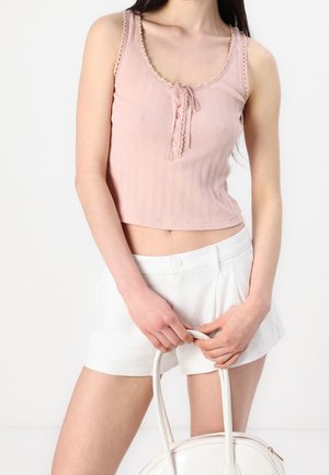 Femme portant un crop top rose sans manches et un short blanc taille haute, tenant un sac à main blanc devant un fond uni.
