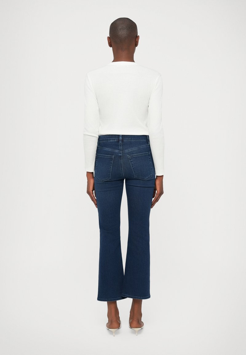 Person wearing dark blue flared jeans, white long-sleeve top, and beige kitten heels, standing facing away against a plain background.