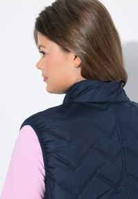 Chaleco acolchado azul marino con un cuello alto, que presenta un patrón en zigzag y un tejido ligero. Llevado sobre una camiseta rosa claro.