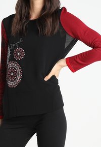 Maglietta nera con maniche lunghe rosse, caratterizzata da eleganti motivi circolari in bianco e bordeaux. Il tessuto appare morbido e leggermente strutturato.