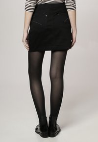 Minifalda de mezclilla negra con acentos metálicos y dos bolsillos traseros, combinada con medias negras transparentes y botas de tobillo. La parte superior a rayas es parcialmente visible.