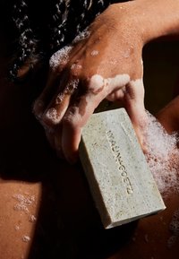 Grüne Seife mit strukturierter Oberfläche und kleinen schwarzen Punkten, in einer Hand mit Blasen gehalten, vor einem braunen, nassen, strukturierten Hintergrund.