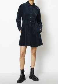 Abito in corduroy blu scuro con chiusura a bottoni, colletto, maniche lunghe e tasche laterali, abbinato a stivaletti neri alla caviglia.