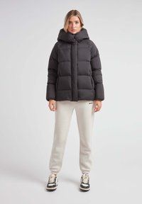 Czarna kurtka puchowa z wysokim kołnierzem i pikowanym wzorem, w połączeniu z jasnymi spodniami dresowymi. Model stoi na neutralnym tle.