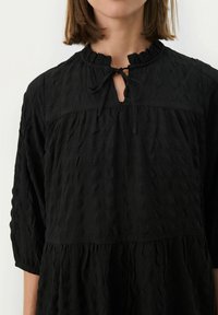 Schwarzes, texturiertes Kleid mit einem Rüschenausschnitt, gebundenem Verschluss und dreiviertelÄrmeln, das eine lockere Passform und ein mehrschichtiges Design aufweist.