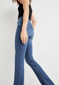 Isik sinistes laienevate teksade ja musta käisteta topiga, kõndimas üksluisel valgel taustal.