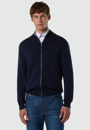 Joven con suéter azul marino con cremallera sobre camisa blanca de cuello y jeans azules, de pie frente a un fondo liso gris claro.