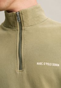 Close-up van een persoon die een olijfgroen ritspulli draagt met de tekst "MARC O'POLO DENIM" op de borst.