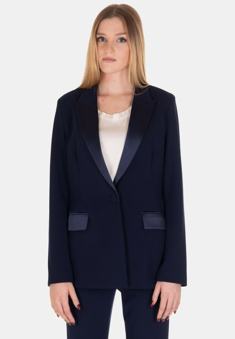 Blazer blu navy con rever in satin e dettagli sulle tasche, vestibilità sartoriale, maniche lunghe e chiusura con un solo bottone. Indossato sopra una maglietta chiara.