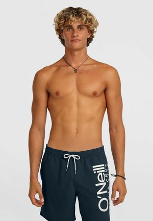 Jonge man met krullend blond haar, gekleed in marineblauwe O'Neill zwembroek, staand tegen een eenvoudige lichte achtergrond, voorwaarts gekeerd.