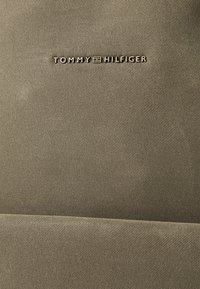 Kahki-stoff med en teksturert overflate som har det pregete logoen "TOMMY HILFIGER" i gull. Enkel, elegant design. Ingen ekstra aksenter.