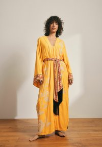 Geltonas kimono stiliaus chalatas su gėlių siuvinėjimu, ilgomis rankovėmis, giliu V formos iškirpimu, pabrėžtas raštuota diržu su kutais.