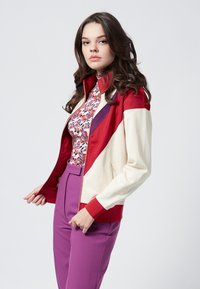 Giacca rossa e crema con zip, colletto alto e blocchi di colore geometrici, abbinata a pantaloni viola su misura e una maglietta floreale.