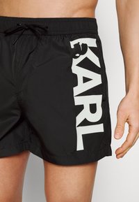 Juodos maudymosi šortai, pagaminti iš lengvos medžiagos, turi didelį baltą "KARL" spaudą šone ir reguliuojamą raištelį juosmens.