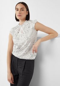 White blouse with black speckled pattern, short ruffled sleeves, keyhole neckline detail, paired with dark trousers. Smooth fabric texture.