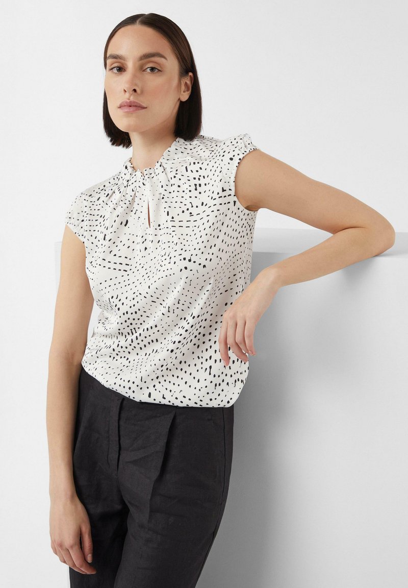 White blouse with black speckled pattern, short ruffled sleeves, keyhole neckline detail, paired with dark trousers. Smooth fabric texture.