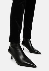 Schwarze Leder-Stiefeletten mit spitzer Zehenform und schlankem, hohen Absatz. Glatte Textur mit minimaler Verzierung, kombiniert mit schwarzen, taillierten Hosen.