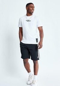 Camiseta blanca con el logo "ELEVEN DEGREES", combinada con pantalones cortos negros con franjas laterales blancas, y zapatillas blancas con tela texturizada.