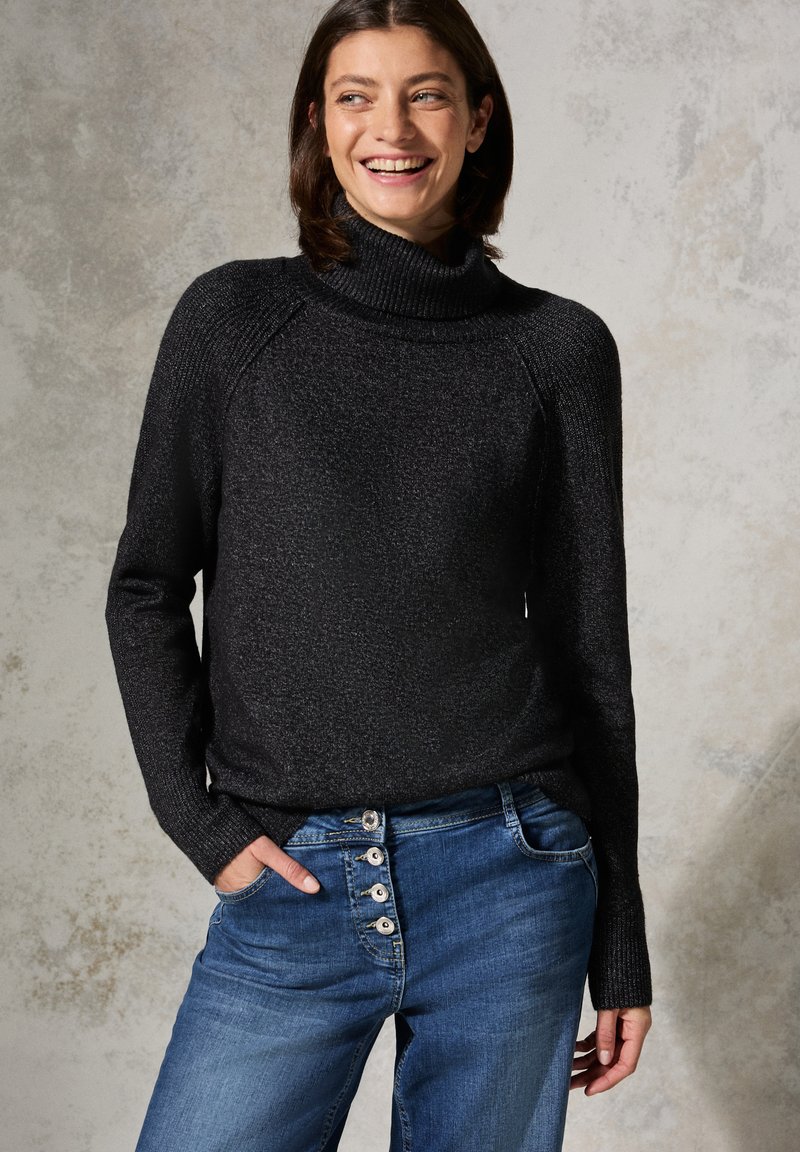 Maglione a collo alto nero con finitura testurizzata e maniche raglan, abbinato a jeans blu con chiusura a bottoni. Modello sorridente, mani in tasca.