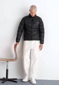 Svart dunjakke med glidelåslomme, quiltet design og høy krage, kombinert med hvite bukser og hvite sneakers. Mot en ensfarget bakgrunn.