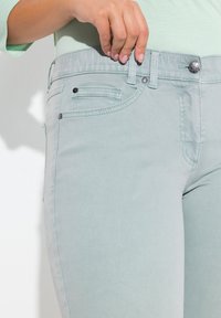 Jeans de mezclilla verde claro con un diseño ajustado, que cuentan con bolsillos frontales, costuras decorativas y un cierre de botón en la cinturilla.