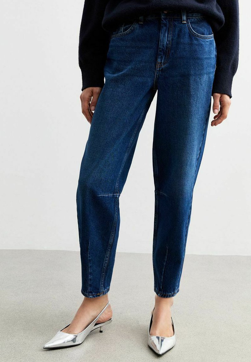 Jeans di jeans scuri a vita alta con gambe diritte, dotati di tasche frontali, cuciture subtle e una superficie liscia. Abbinati a scarpe argentate a punta.