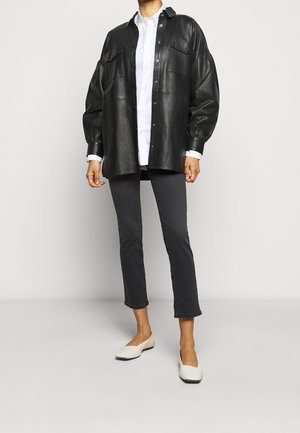 Musta nahkainen tilava paitatakki, jossa napitukset, alla valkoinen kauluspaita, harmaat kapenevat housut ja kermaiset slip-on-kengät.