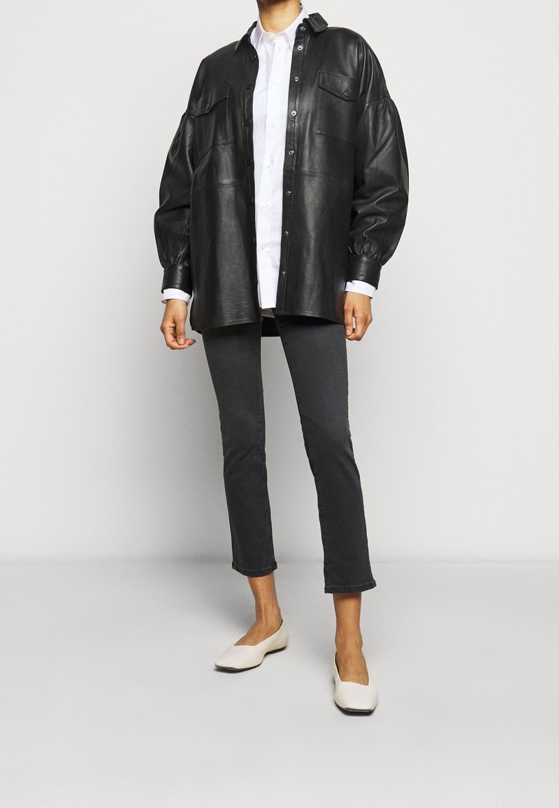 Chemise-veston surdimensionnée en cuir noir avec fermeture à boutons, chemise blanche à col en dessous, pantalon gris fuselé et chaussures slip-on crème.