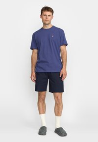 Navy blue cotton t-shirt with short sleeves and a small logo on the chest, paired with navy shorts, white socks, and grey clogs.
