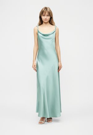 Ung kvinde stående iført en gulvlængde, lyseblå satin kjole med tynde stropper og hvide hælede sandaler på en ensfarvet baggrund.