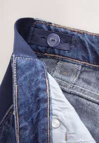 Denim linning med mørkeblåt stof, med en blå knap og lys blå inderforing. Synlige syninger og teksturdetaljer.