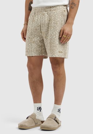 Beige korte broek met patroon, gedragen met witte sokken met zwarte letters en beige instapschoenen met gespen op een lichte achtergrond.