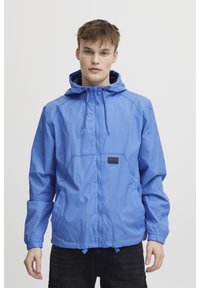 Jachetă ușoară bleu cu glugă, fermoar frontal, șnururi ajustabile și o mică insignă logo. Textură netedă și design casual.