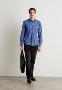 Cămașă albastră cu mânecă lungă, cu închidere cu nasturi și un accent roșu, combinată cu pantaloni negri și purtând o geantă de tote neagră. Papuci plata la picioare.