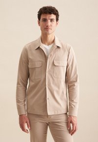 Lys beige skjorte med lange ærmer, to brystlommer og spids krave, lavet af et glat stof med en mat finish.