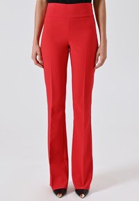 Donna che indossa pantaloni rossi a vita alta con pieghe davanti, abbinati a tacchi neri a punta con punte dorate.