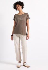 Camiseta verde oliva de manga corta, con escote en forma de barco y diseño impreso de "NATURAL". Combinada con pantalones sueltos de color beige claro y zapatillas blancas.